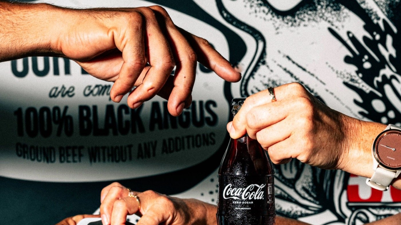 Hands opening a Coca-Cola bottle in front of a graffiti wall